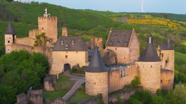 bourscheid castle ch&acirc;teau de green valley cover hills behind walls towers exterior structure visitor parking windmill atop to left right up close circling drone 