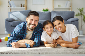 Happy family parents and child smiling together. They lie on a soft carpet, relaxing and bonding in...