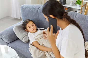 Sick child with mother on sofa. A parent calls the doctor, describing fever and virus illness while...