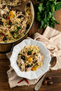 Overhead view of a hearty Mediterranean chicken and orzo pasta dish with olives, lemon, and fresh parsley, served in a bowl and a cast iron pan on a rustic wooden table.