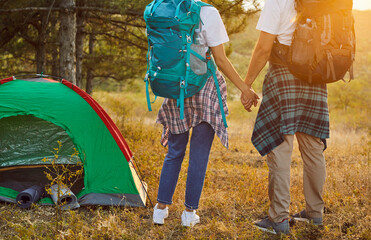 Couple hikers with backpacks holding hands, tourists standing by tent at sunset. People enjoying...