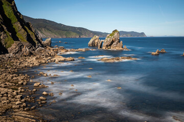 Dramatic coastal seascape with ancient rock formations and ethereal blurred ocean waves under a clear blue sky