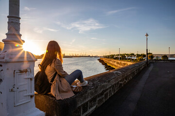 Young woman contemplates a beautiful coastal sunset over a busy harbor with industrial cranes...