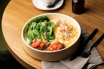 Gourmet breakfast bowl with creamy scrambled eggs, succulent shrimp, fresh green salad, and tomato bruschetta on a wooden table.