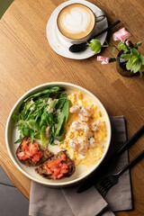 A gourmet breakfast or brunch spread: creamy scrambled eggs with shrimp, fresh green salad, and tomato bruschetta, served alongside a latte on a warm wooden table. Inviting, healthy meal.