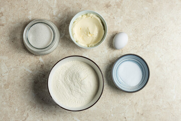  Top-view shot of baking ingredients on a marble surface: sugar in a jar, butter in a bowl, an egg, flour in a large bowl, and a small bowl of salt. Clean, minimalist food styling. Ideal for recipe bl