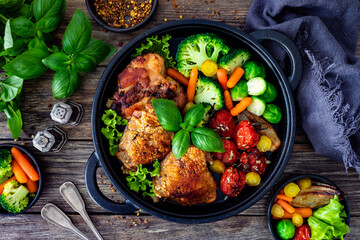 Roasted chicken thighs and vegetables in cast iron pan on wooden table. Top view