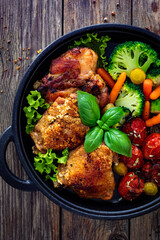 Roasted chicken thighs and vegetables in cast iron pan on wooden table. Top view