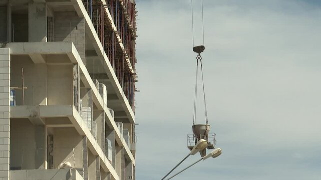 Crane lifts a concrete hopper alongside a multi-story building under construction. Workers are busy on scaffolding, building the new high-rise structure against a clear blue sky.