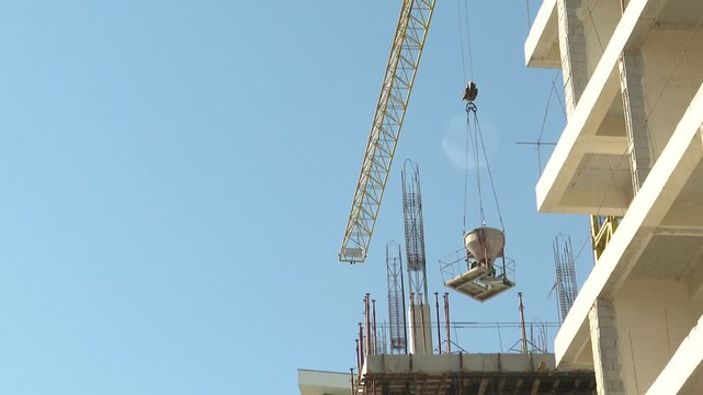 Crane lifts a concrete hopper alongside a multi-story building under construction. Workers are busy on scaffolding, building the new high-rise structure against a clear blue sky.