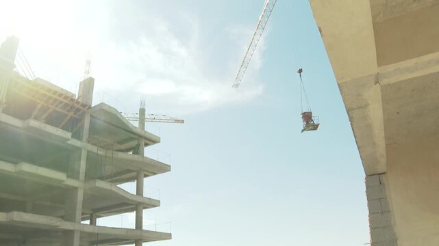 Crane lifts a concrete hopper alongside a multi-story building under construction. Workers are busy on scaffolding, building the new high-rise structure against a clear blue sky.