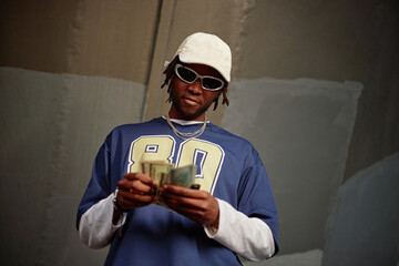 Black young adult man wearing sunglasses and cap counting cash while standing against painted wall,...