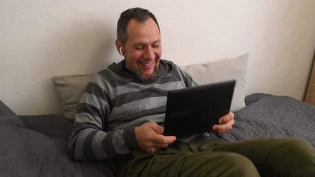 A bearded middle-aged man sits on a bed in the bedroom watching football, rooting for a sports team, the fan is upset about the loss and rejoices at the success