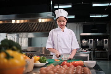 One young trainee chef girl in white apron is slicing carrot and vegetable spice ingredients with knife, preparing a delicious meal in restaurant kitchen, intern gourmet foods, and cooking culinary.