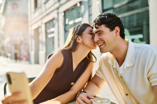 Young couple enjoying conversation and coffee together and taking a selfie with a smartphone at an outdoor urban cafe table - Powered by Adobe