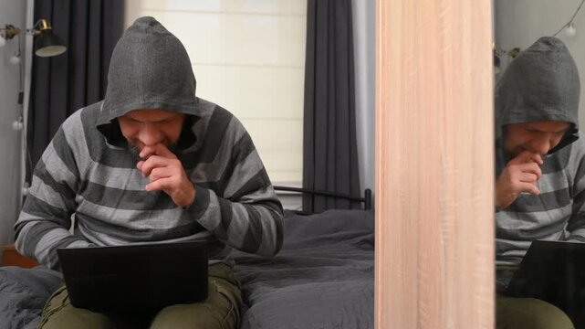 A bearded middle-aged man sits on a bed in the bedroom watching football, rooting for a sports team, the fan is upset about the loss and rejoices at the success