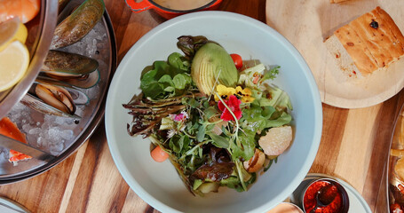 Fresh avocado salad with greens, edible flowers, and cherry tomatoes served at rustic restaurant. Includes seafood and bread on wooden table.