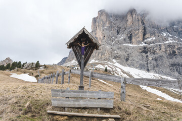 Wooden crucifix with bench on alpine hiking trail in Dolomites, Italy, with snowy Sassolungo mountain peak in background.