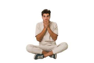 Young caucasian man sitting on the floor isolated on white background shocked, covering mouth with hands, anxious to discover something new.