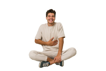 Young caucasian man sitting on the floor isolated on white background touches tummy, smiles gently,...