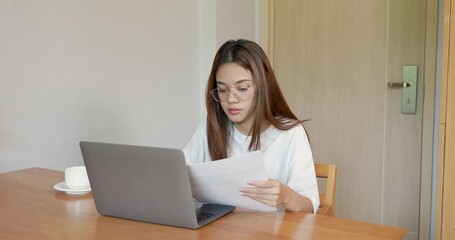 Asian adult woman reads printed document beside laptop at home desk with serious face during long remote work session, feeling tired and focused while processing multiple tasks under pressure - Powered by Adobe
