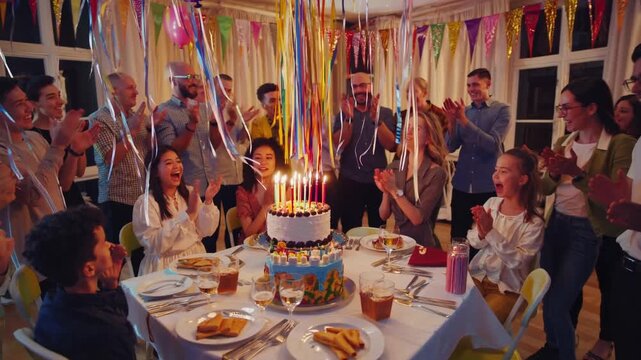 Large multi generational family and friends clapping and singing around a birthday cake with lit candles. Joyful atmosphere with balloons