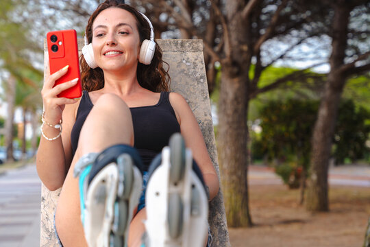 Smiling young woman with inline skates sitting outdoors, listening to music with headphones and holding red smartphone. Lifestyle, sport, technology, fitness, leisure, summer, fun, urban freedom and - Powered by Adobe