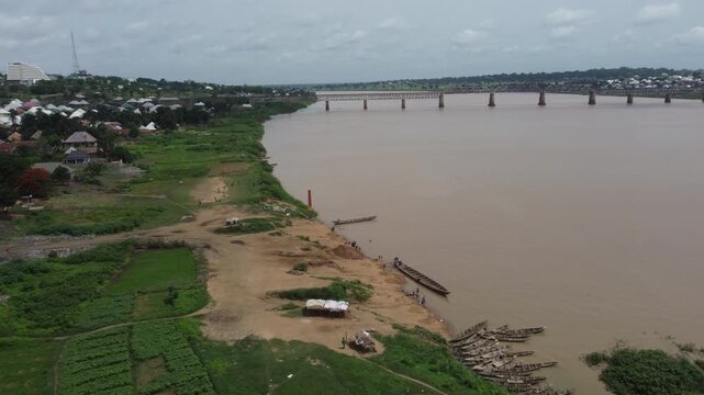 Makurdi, Nigeria - 13 October 2025: Aerial view of boats lining the sandy bank of the Benue River near lush greenery, with a bridge stretching across.