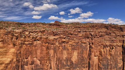 Aerial view of red rock cliffs rise dramatically against a bright blue sky dotted with fluffy white clouds, creating a stark contrast of colors and textures, Moab, Utah, United States.