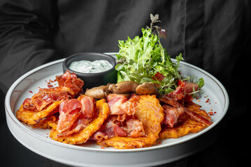 Brunch plate with crispy bacon, hash browns, sautéed mushrooms, fresh salad, and creamy herb sauce. Held by person in dark outfit. Rustic, hearty, and appetizing presentation.