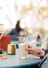 Hand holding a refreshing drink at outdoor café table.Close-up of a person holding a glass of cold drink with foam at an outdoor café table on a sunny day. Relaxed atmosphere and warm natural light, 
