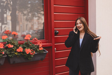 Cheerful young woman talking on her smartphone near a bright red door with flowers. Warm urban lifestyle moment showing communication, positivity and stylish casual fashion in natural daylight. Wow!