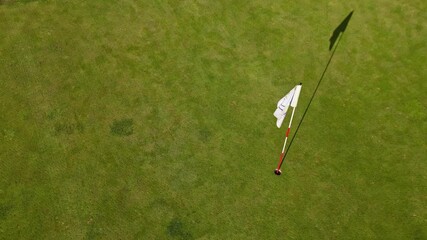 Descending camera over well-maintained golf course green, revealing a white flagstick casting long shadow across the smooth grass, begining elevated and gradually lowers toward the putting surface