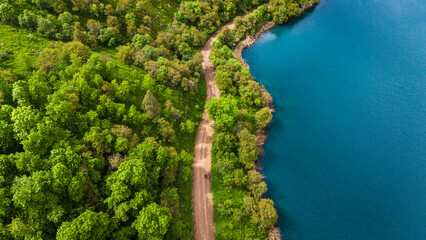 Aerial view of a dirt road running along a turquoise lake surrounded by lush green forest. Scenic natural landscape captured from drone in summer daylight