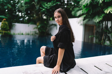 Elegant woman sits by pool looking calmly at camera, exuding digital confidence, poised energy and independence in modern smart lifestyle.
