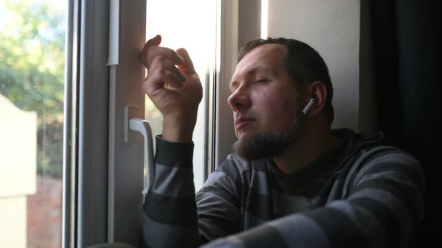 A bearded man listens to music while sitting by the window, sad, crying, loneliness, autumn depression