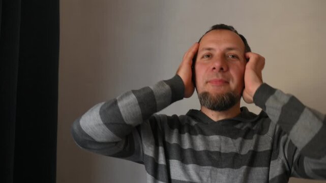 A bearded man listens to music on a portable speaker at home and dances