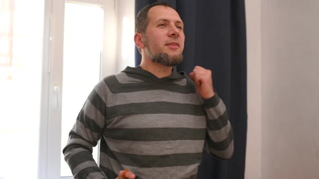 A bearded man listens to music on a portable speaker at home and dances