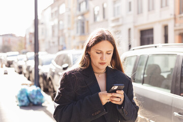 Young woman in oversized blazer using smartphone on a bright urban street. Natural lifestyle moment with warm sunlight, modern style and city atmosphere, ideal for tech, fashion or lifestyle concepts.