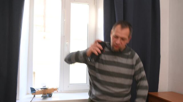 A bearded man listens to music on a portable speaker at home and dances