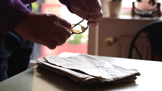 Closeup of a man's hands taking glasses from a pile of newspapers on a kitchen table to read them