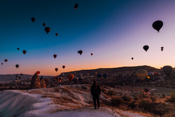 Goreme, Turkiye - 10 October 2019: View of a lone figure stands silhouetted against the soft glow of dawn as colorful hot air balloons drift serenely above the otherworldly landscape.