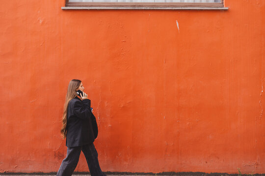 Serious young adult business woman walks and talks on phone, solves problems, holds documents, notebook, tablet and bag on background of orange wall. Girl dressed in black jacket, denim jeans. Side. - Powered by Adobe