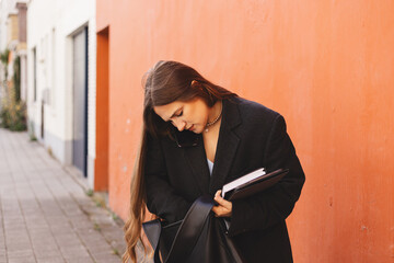 Young businesswoman multitasking while talking on smartphone, searching in bag and holding planner....