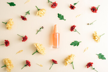 Glass cosmetic bottle with yellow and red chrysanthemums on beige background. Flat lay natural beauty composition. Top view, mockup