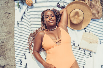 Top-down view of young woman with braided hair reading notebook on towel next to sunhat, phone, and...