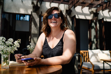Confident middle-aged woman smiling at camera while seated with smartphone and journal, radiating positivity, digital fluency and entrepreneurial spirit in relaxed setting.