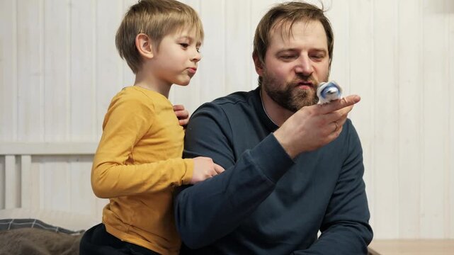 Father and son enjoying at home. Young boy and his daddy playing with funny pet budgerigar parrot. Happy family. Tamed budgie parakeet. People take care of and play with bird. Cute domestic animals