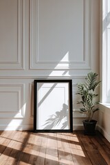 Fototapeta premium Black frame on a paneled white wall with a potted plant, bathed in sunlight