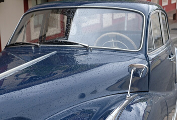 Hood and windshield of vintage dark blue car, covered in fresh raindrops. Chrome trim, side mirror, and the view through wet glass, revealing vintage steering wheel inside. Vintage car design.